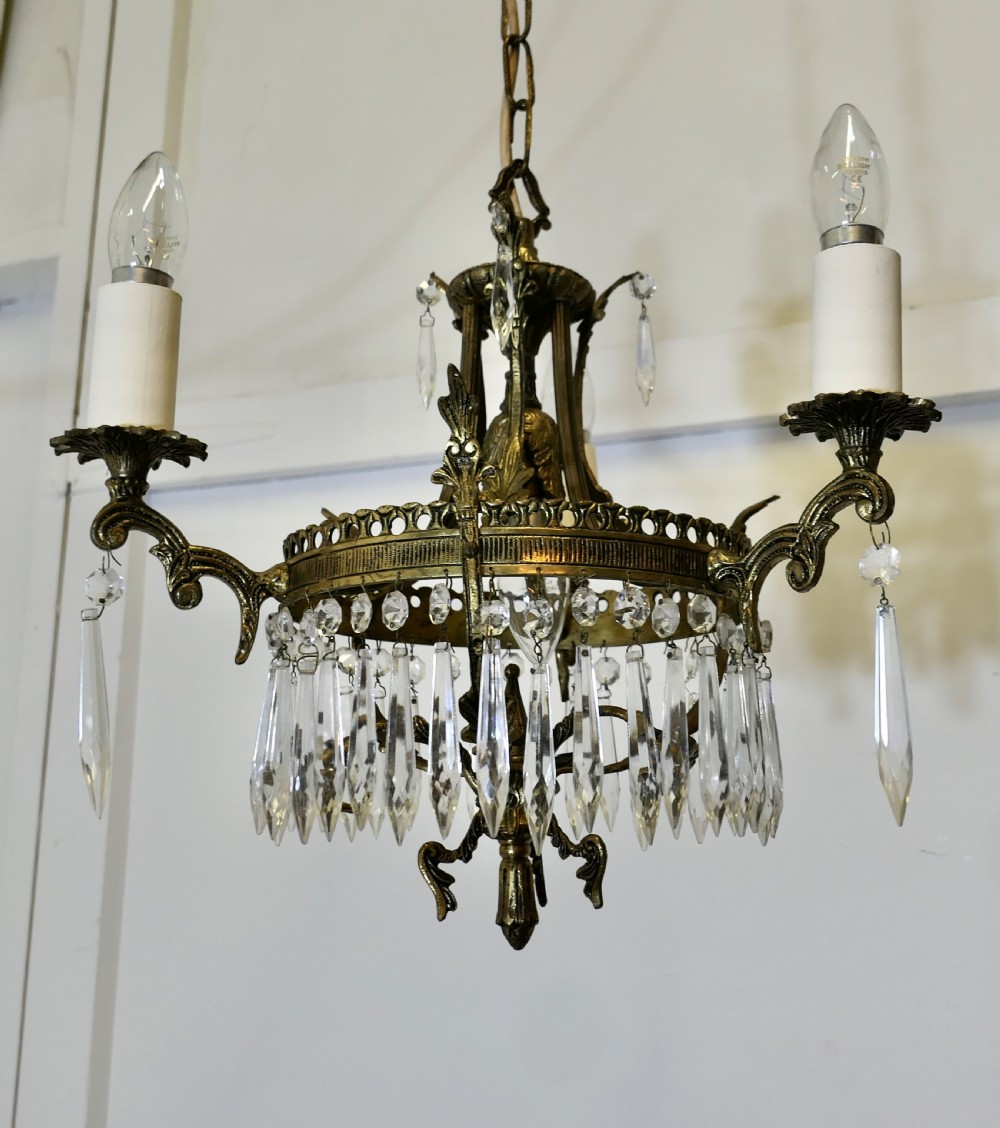A three-arm brass chandelier adorned with cut crystal glass droplets and candle sleeves hanging from a brass chain suspension. View IV