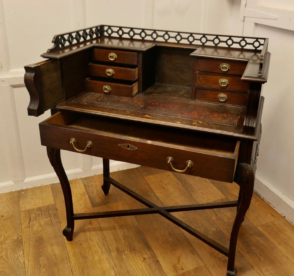 An antique dark mahogany writing desk featuring an openwork fretwork gallery, inset leather writing surface, multiple small drawers, and cabriole legs supported by an X-stretcher. View VI