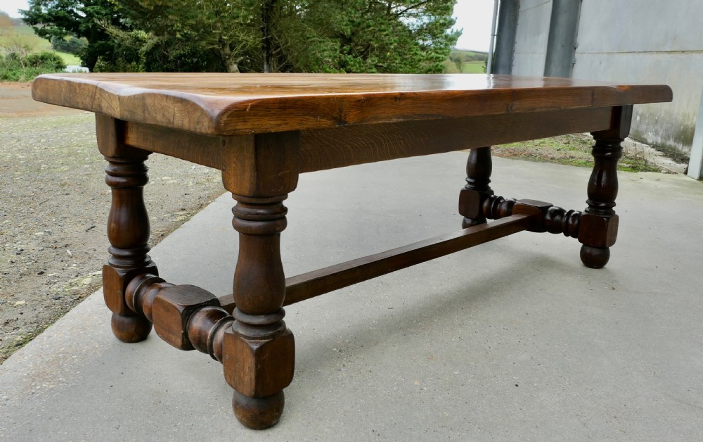 A large solid oak refectory dining table featuring a thick plank top, heavy turned baluster legs, and a low central stretcher base. View V