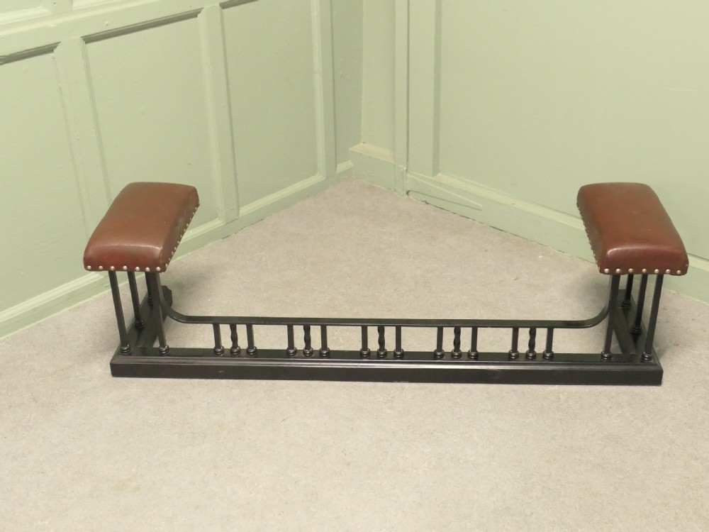 An Art Deco style club fender featuring a black iron frame with twisted vertical spindles and two elevated tan leather padded seats trimmed with studs.