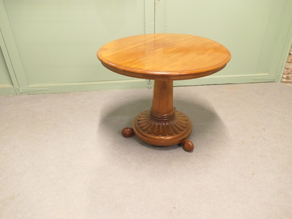A circular cherrywood centre table featuring a polished tilt-top surface, a central stem, and a carved petal-form pedestal base on ball feet. View VII