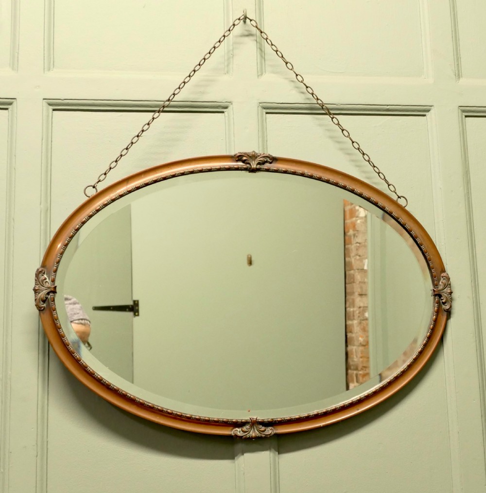 An oval copper-framed wall mirror featuring decorative acanthus leaf mounts at four points and a beaded inner border, suspended by a metal chain. View II