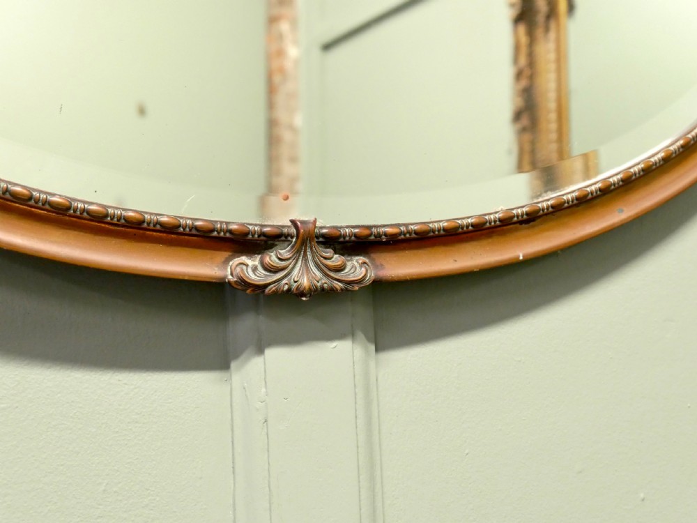 An oval copper-framed wall mirror featuring decorative acanthus leaf mounts at four points and a beaded inner border, suspended by a metal chain. View IV