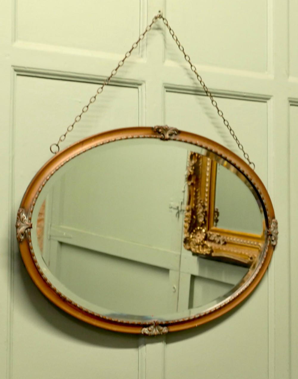 An oval copper-framed wall mirror featuring decorative acanthus leaf mounts at four points and a beaded inner border, suspended by a metal chain. View V