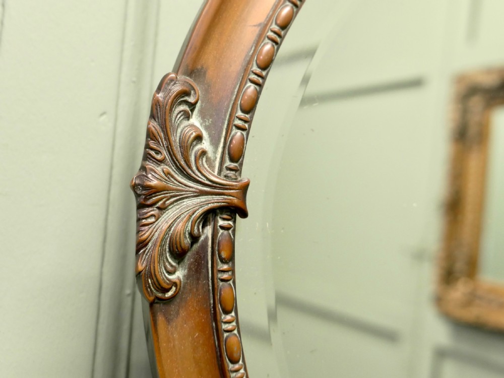 An oval copper-framed wall mirror featuring decorative acanthus leaf mounts at four points and a beaded inner border, suspended by a metal chain. View VI
