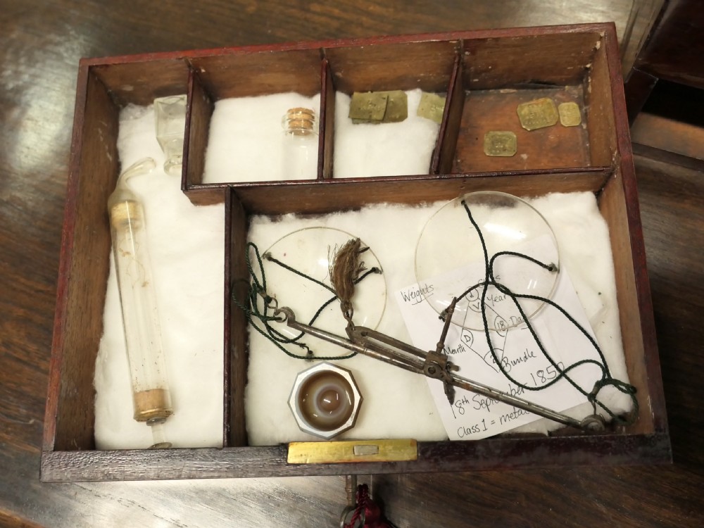 An open antique mahogany apothecary chest featuring velvet-lined compartments filled with glass bottles and a lower drawer containing scales. View V