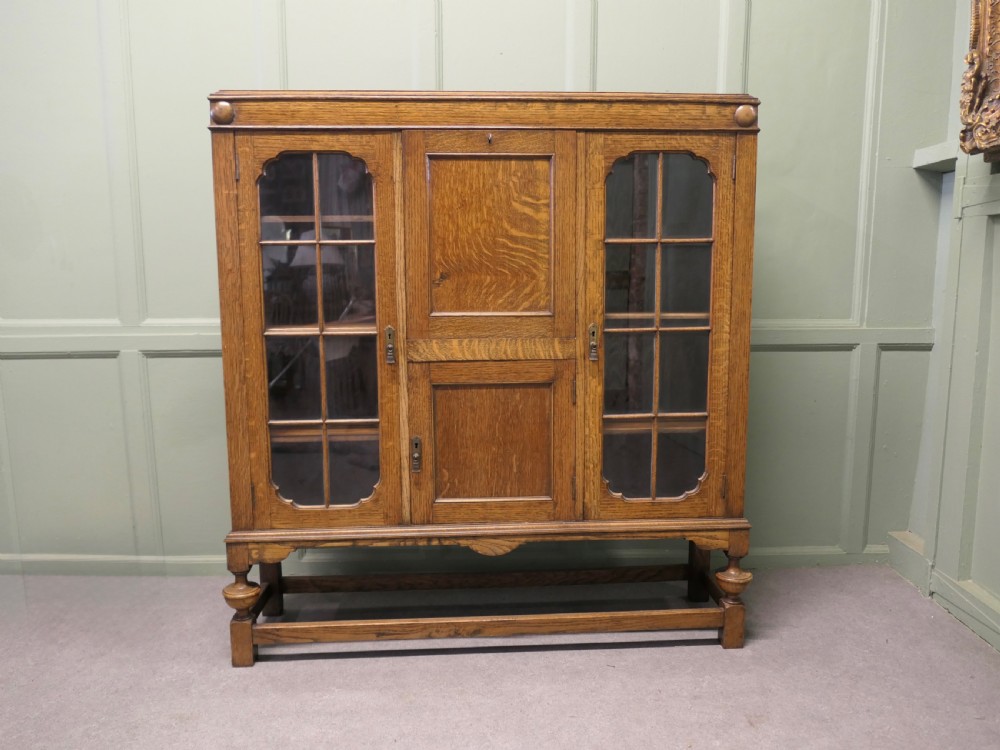 An Arts and Crafts golden oak bureau cabinet featuring twin side glazed display compartments flanking a central fall-front desk flap and cupboard base.