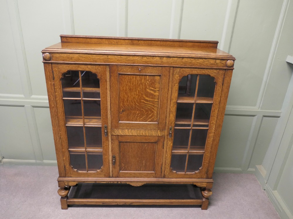 An Arts and Crafts golden oak bureau cabinet featuring twin side glazed display compartments flanking a central fall-front desk flap and cupboard base. View II