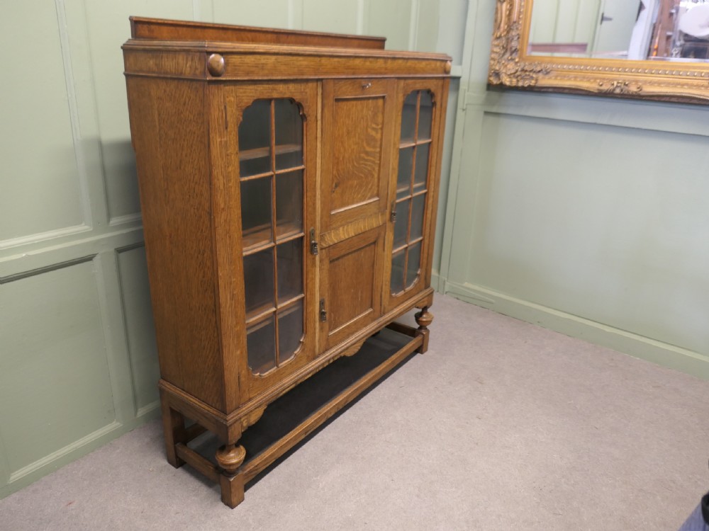 An Arts and Crafts golden oak bureau cabinet featuring twin side glazed display compartments flanking a central fall-front desk flap and cupboard base. View III