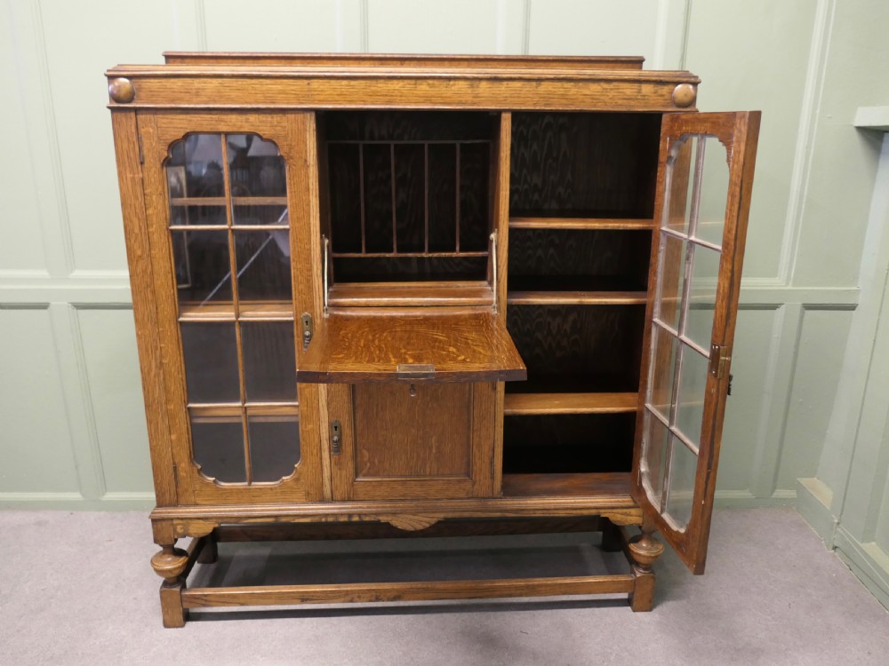 An Arts and Crafts golden oak bureau cabinet featuring twin side glazed display compartments flanking a central fall-front desk flap and cupboard base. View IV