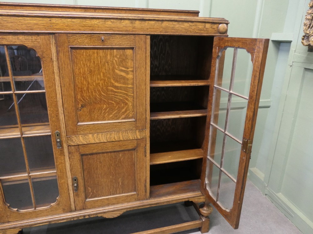 An Arts and Crafts golden oak bureau cabinet featuring twin side glazed display compartments flanking a central fall-front desk flap and cupboard base. View V
