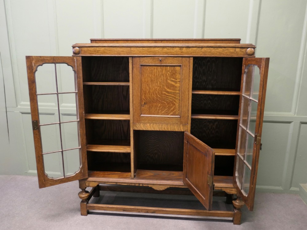 An Arts and Crafts golden oak bureau cabinet featuring twin side glazed display compartments flanking a central fall-front desk flap and cupboard base. View VI