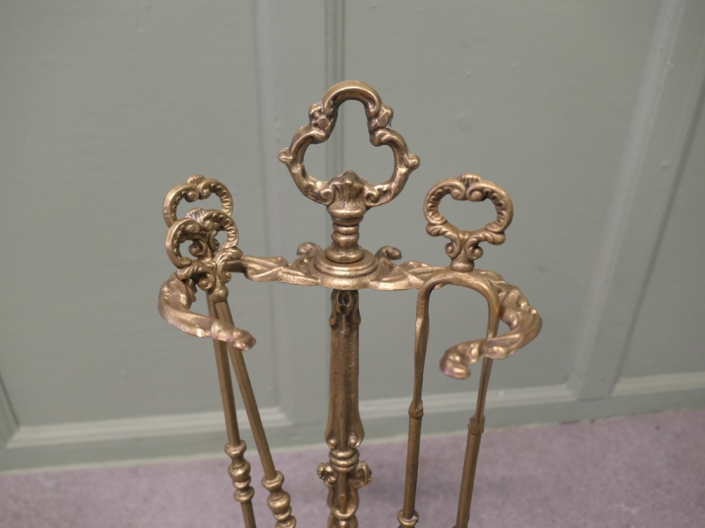 A solid brass Rococo style fireside companion set featuring an ornate scrollwork stand holding a shovel, poker, and tongs. View III