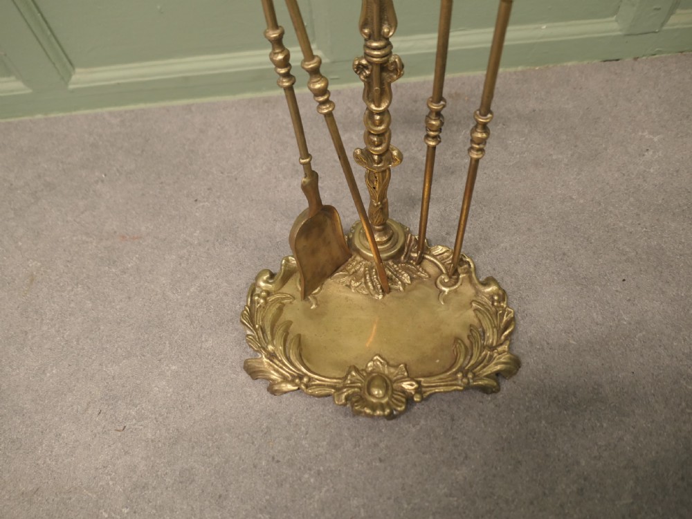A solid brass Rococo style fireside companion set featuring an ornate scrollwork stand holding a shovel, poker, and tongs. View V