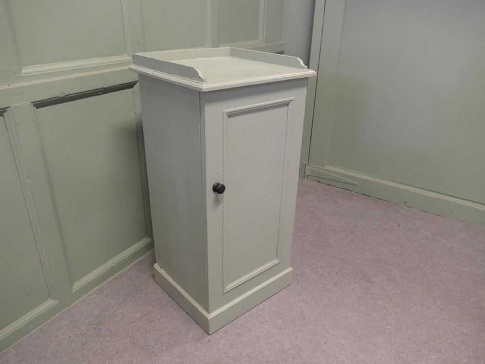 A painted light grey pine bedside cupboard featuring a gallery top rim, a single panelled door with a dark turned knob, and a solid plinth base. View V