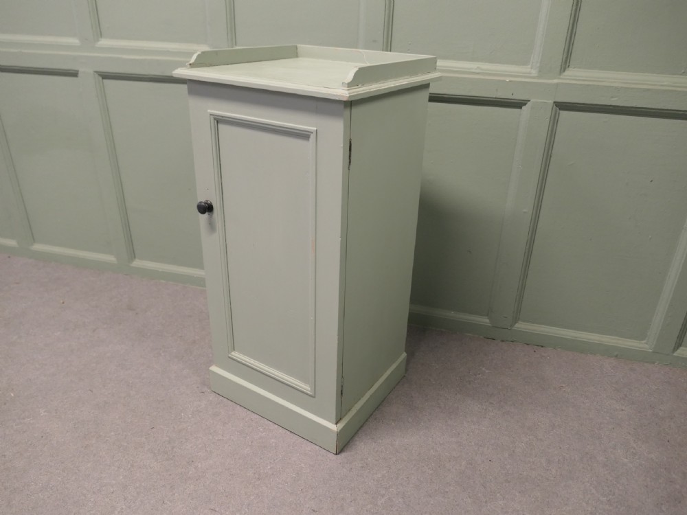 A painted light grey pine bedside cupboard featuring a gallery top rim, a single panelled door with a dark turned knob, and a solid plinth base. View VI