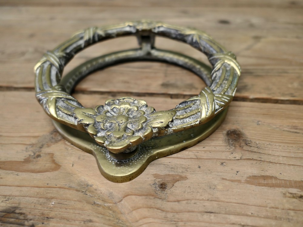 An aged cast brass ring door knocker featuring a floral rosette motif and a textured circular ring.