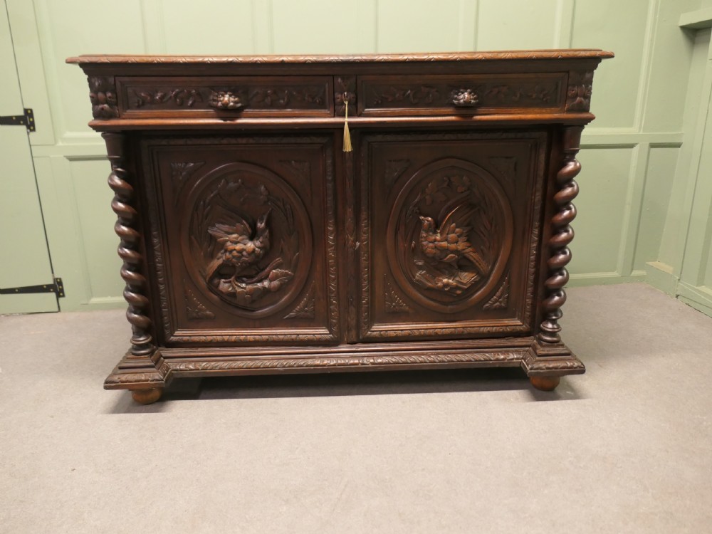 A dark brown carved oak French sideboard featuring two frieze drawers above twin cupboard doors with oval medallions depicting relief-carved game birds, flanked by barley twist columns.