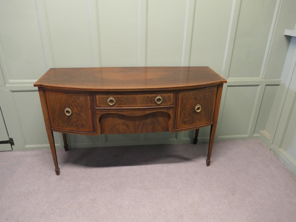 A Regency flame mahogany bowfront sideboard featuring inlaid stringing, a central drawer, two side cupboards, and tapered square legs with brass ring handles. View II