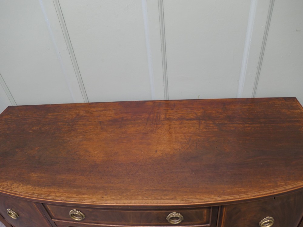 A Regency flame mahogany bowfront sideboard featuring inlaid stringing, a central drawer, two side cupboards, and tapered square legs with brass ring handles. View IV