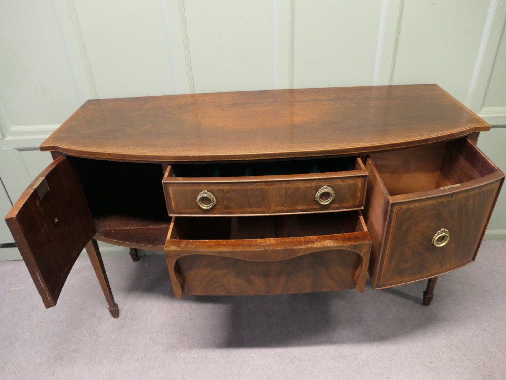A Regency flame mahogany bowfront sideboard featuring inlaid stringing, a central drawer, two side cupboards, and tapered square legs with brass ring handles. View V