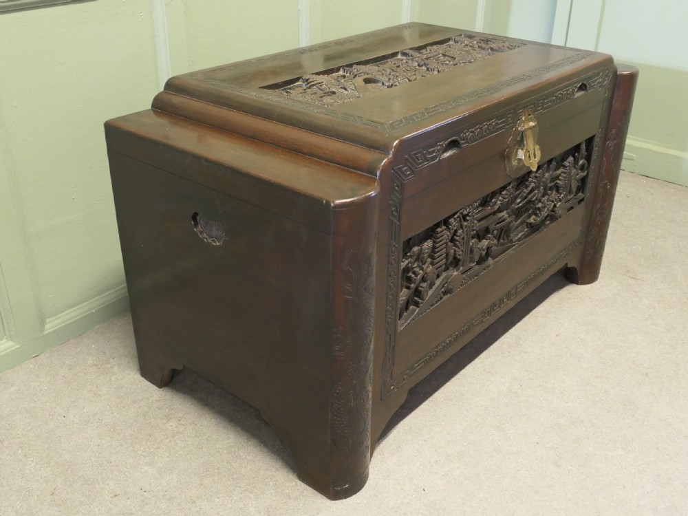 A dark brown carved camphor wood chest featuring detailed relief-carved architectural panels, a stepped Art Deco profile, and a brass latch plate.