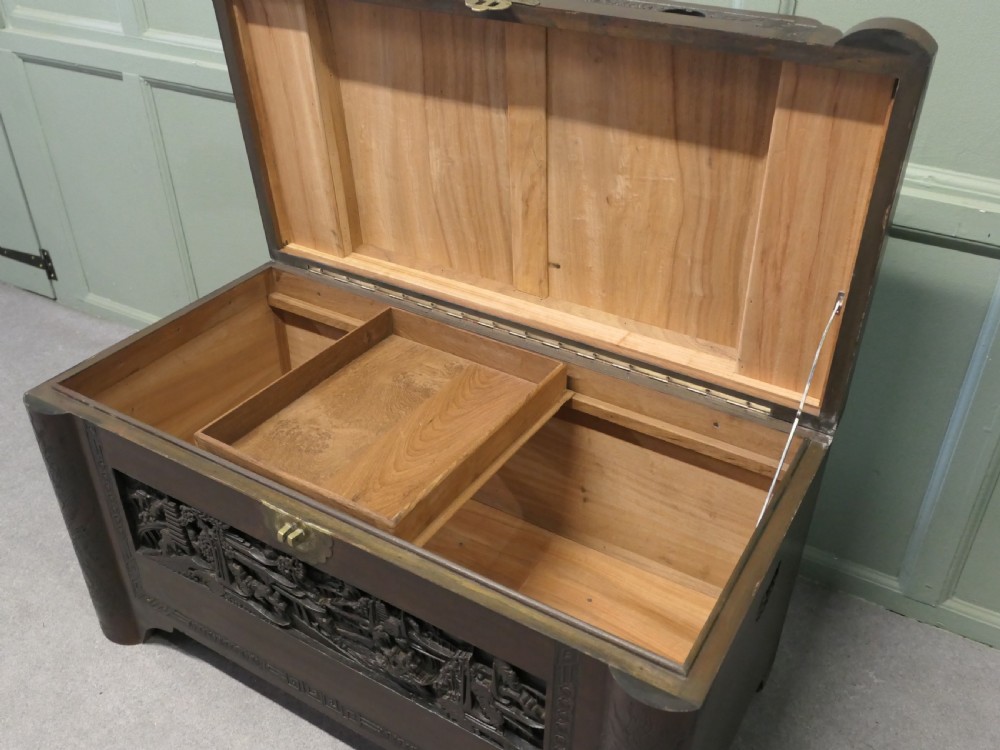 A dark brown carved camphor wood chest featuring detailed relief-carved architectural panels, a stepped Art Deco profile, and a brass latch plate. View III