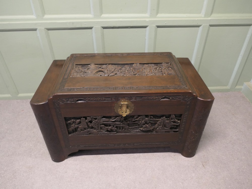 A dark brown carved camphor wood chest featuring detailed relief-carved architectural panels, a stepped Art Deco profile, and a brass latch plate. View V