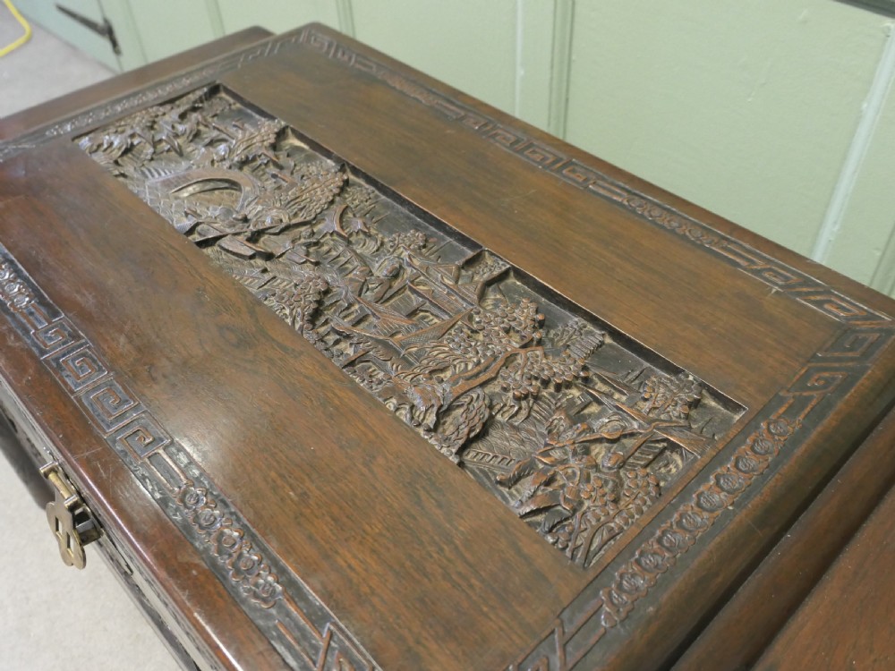 A dark brown carved camphor wood chest featuring detailed relief-carved architectural panels, a stepped Art Deco profile, and a brass latch plate. View VI