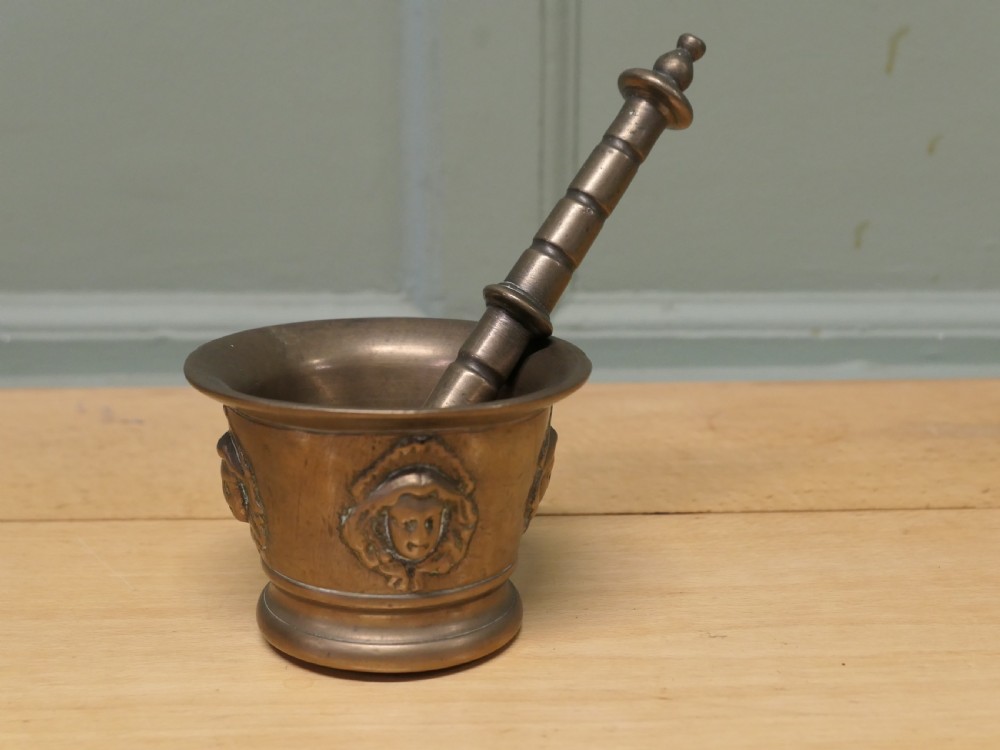 An antique heavy bronze mortar featuring cast relief face medallions on the outer bowl, accompanied by a turned metal pestle resting inside.