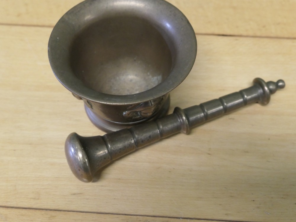 An antique heavy bronze mortar featuring cast relief face medallions on the outer bowl, accompanied by a turned metal pestle resting inside. View II