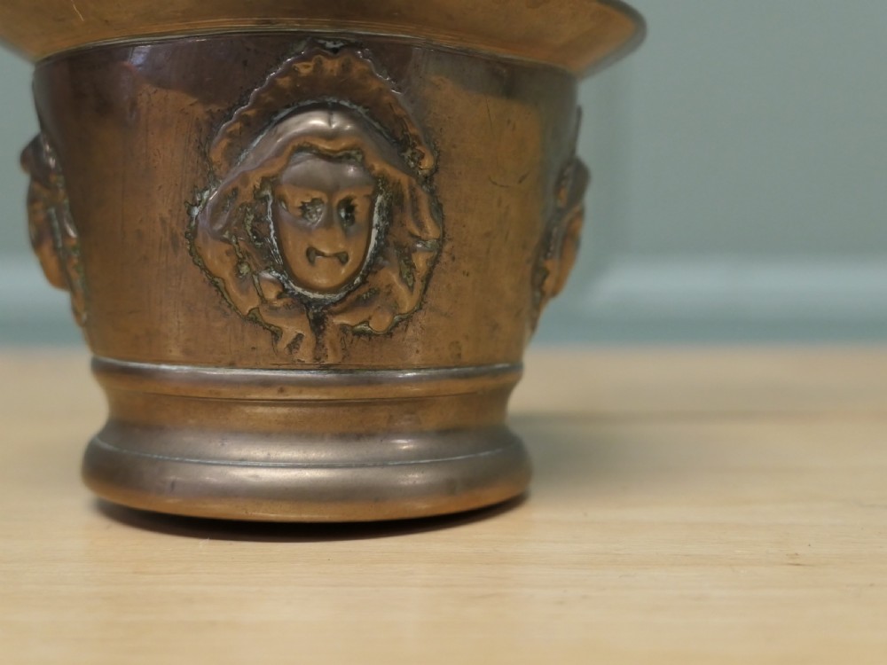 An antique heavy bronze mortar featuring cast relief face medallions on the outer bowl, accompanied by a turned metal pestle resting inside. View III