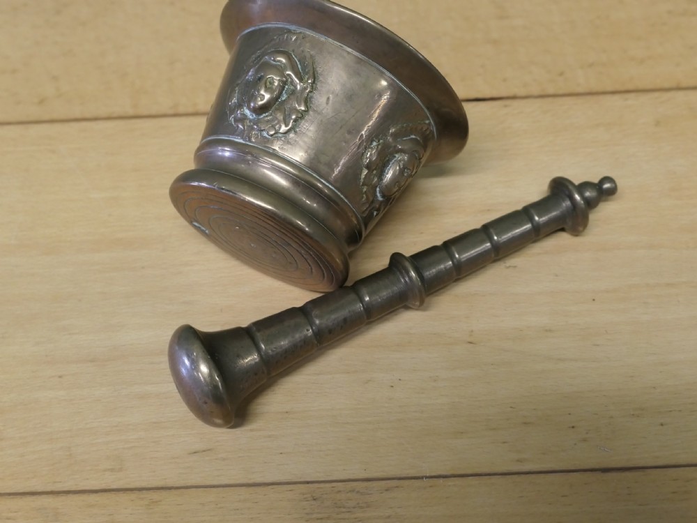 An antique heavy bronze mortar featuring cast relief face medallions on the outer bowl, accompanied by a turned metal pestle resting inside. View IV