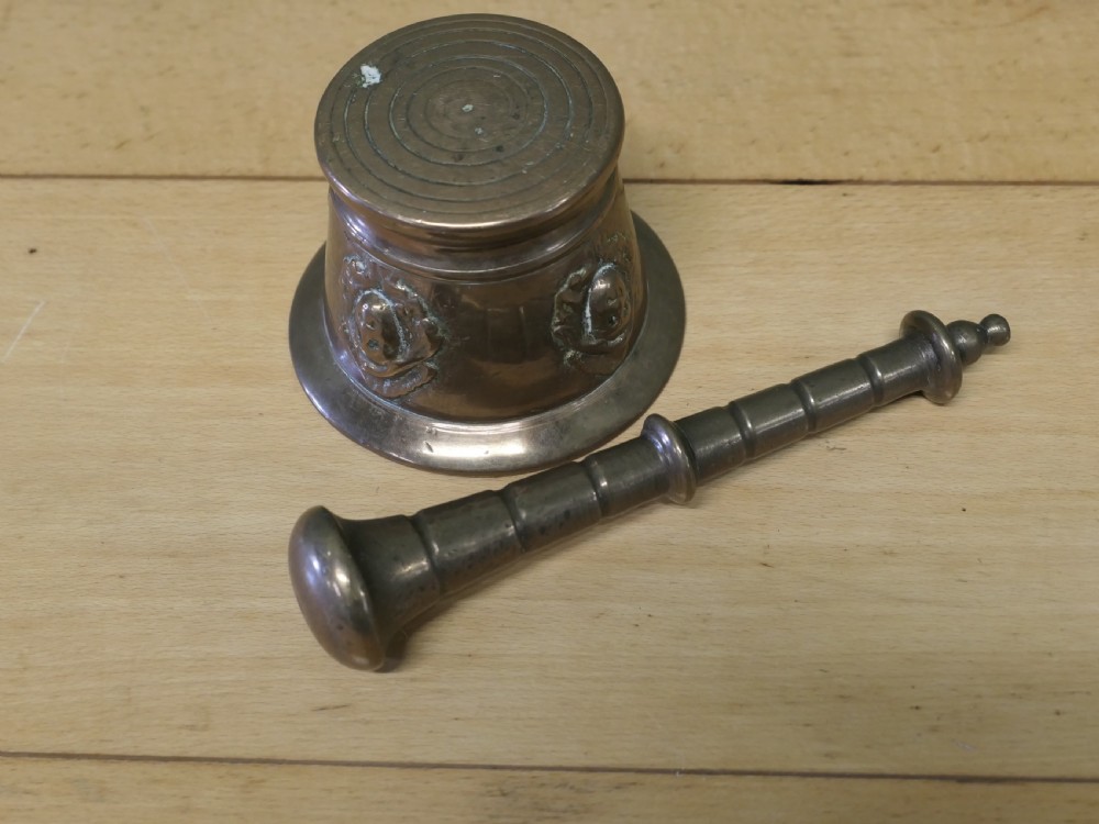 An antique heavy bronze mortar featuring cast relief face medallions on the outer bowl, accompanied by a turned metal pestle resting inside. View V