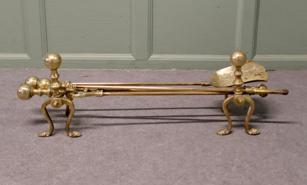 A solid brass fireplace tool set including a shovel, tongs, and poker resting horizontally on matching brass rests.