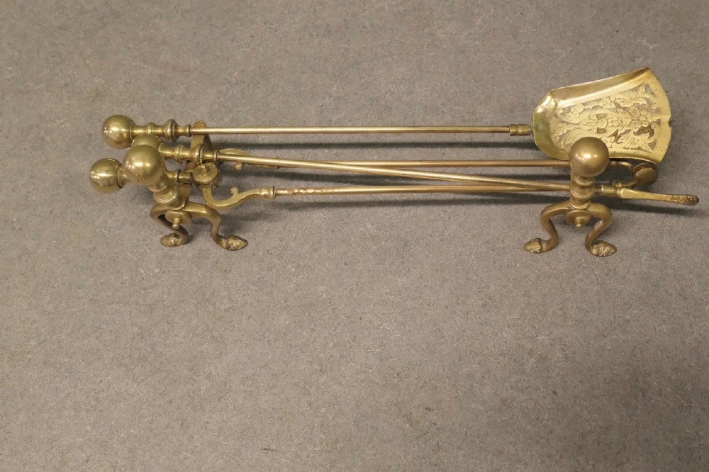 A solid brass fireplace tool set including a shovel, tongs, and poker resting horizontally on matching brass rests. View II