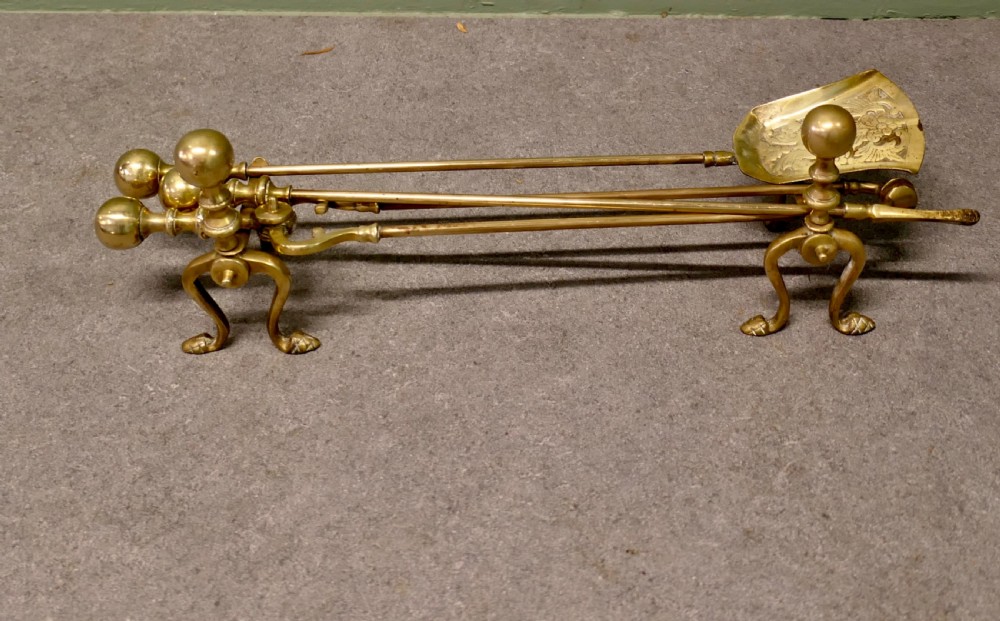 A solid brass fireplace tool set including a shovel, tongs, and poker resting horizontally on matching brass rests. View V