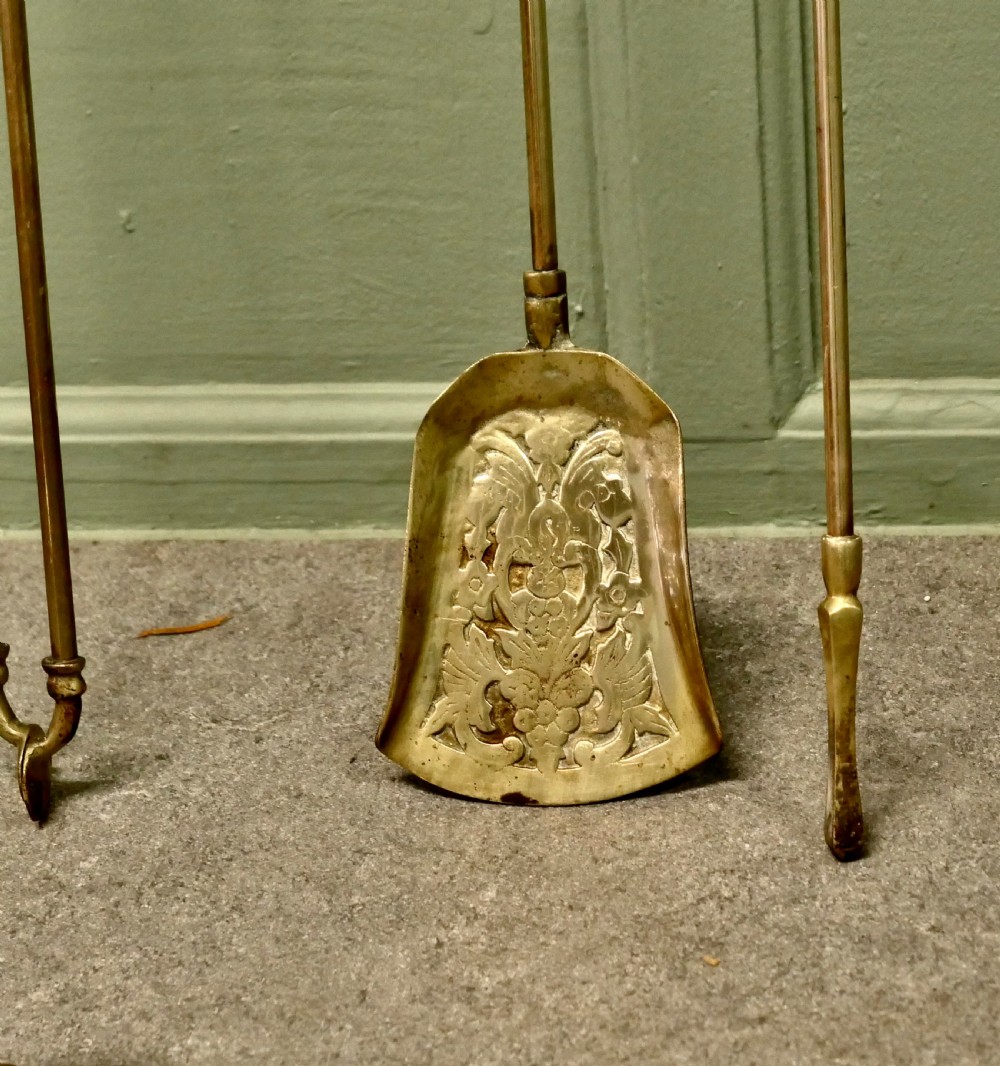 A solid brass fireplace tool set including a shovel, tongs, and poker resting horizontally on matching brass rests. View VI