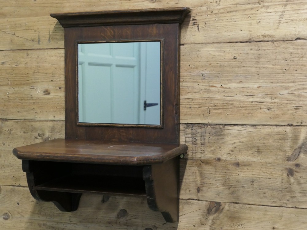 A dark oak wall-mounted mirror featuring a square glass panel surrounded by a moulded wood frame above an integrated display shelf with shaped support brackets.