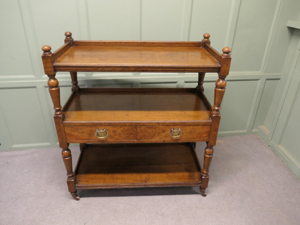 A Victorian oak three-tier buffet featuring turned upright columns, corner finials, two drawers with brass drop handles, and brass castors.