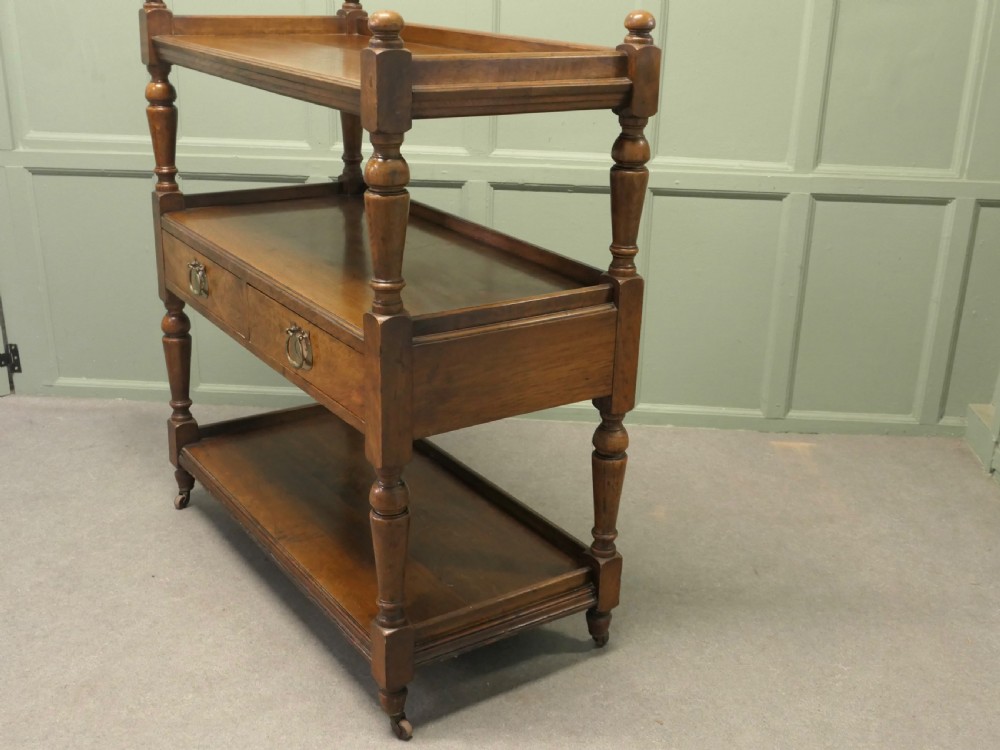 A Victorian oak three-tier buffet featuring turned upright columns, corner finials, two drawers with brass drop handles, and brass castors. View IV