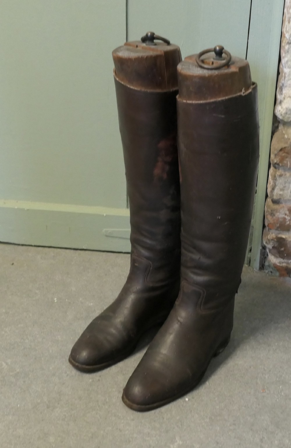 A pair of tall dark brown leather riding boots fitted with carved wooden boot trees featuring metal pull rings.