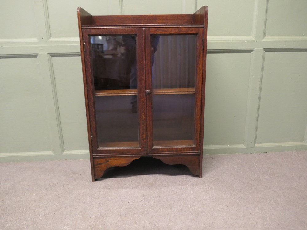 A small dark oak glazed bookcase featuring twin glass panel doors, a raised top gallery, and a shaped lower apron.