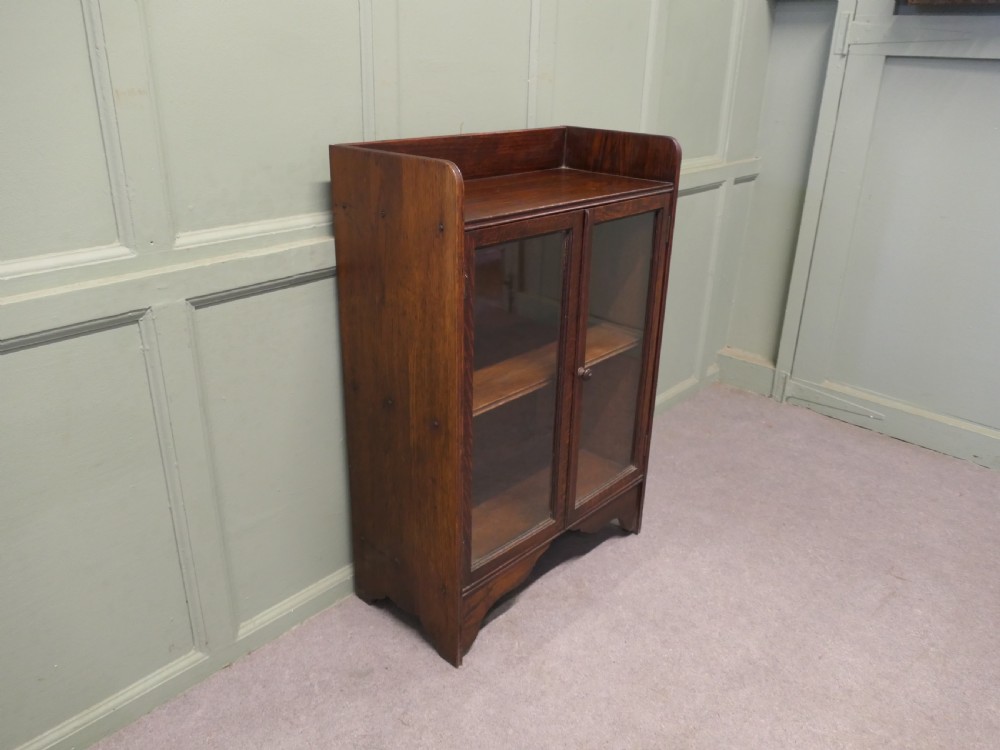 A small dark oak glazed bookcase featuring twin glass panel doors, a raised top gallery, and a shaped lower apron. View II