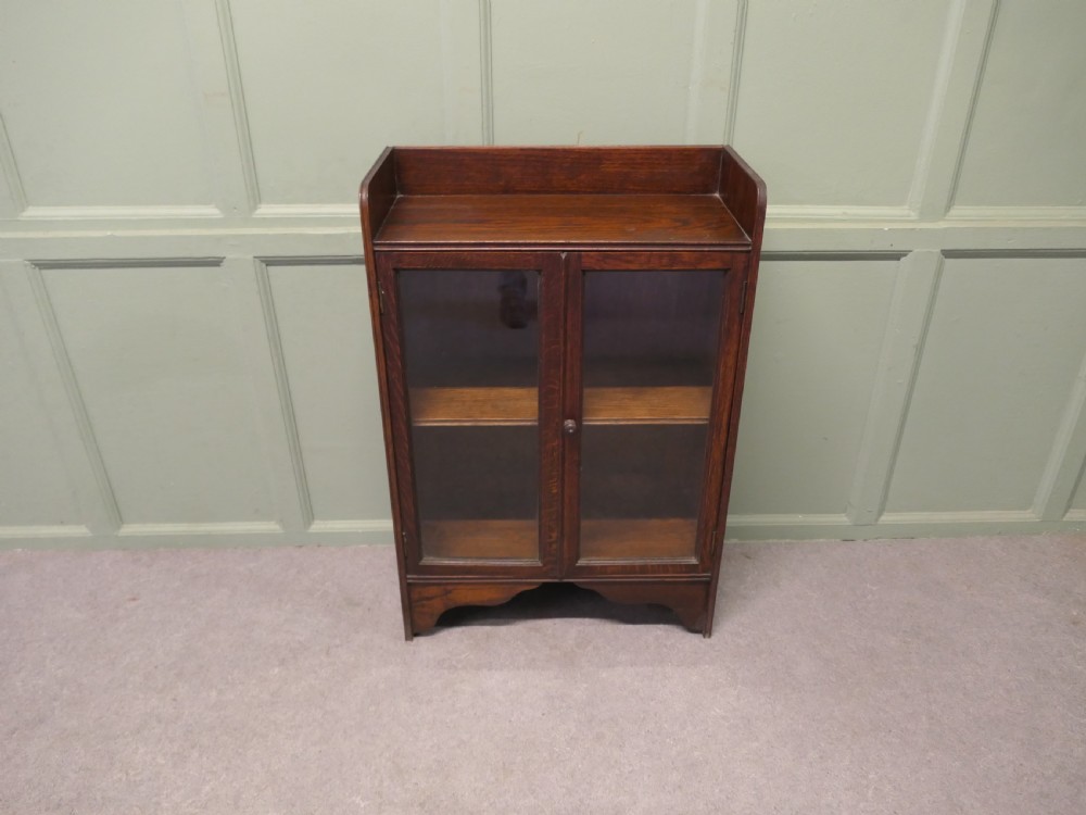 A small dark oak glazed bookcase featuring twin glass panel doors, a raised top gallery, and a shaped lower apron. View III