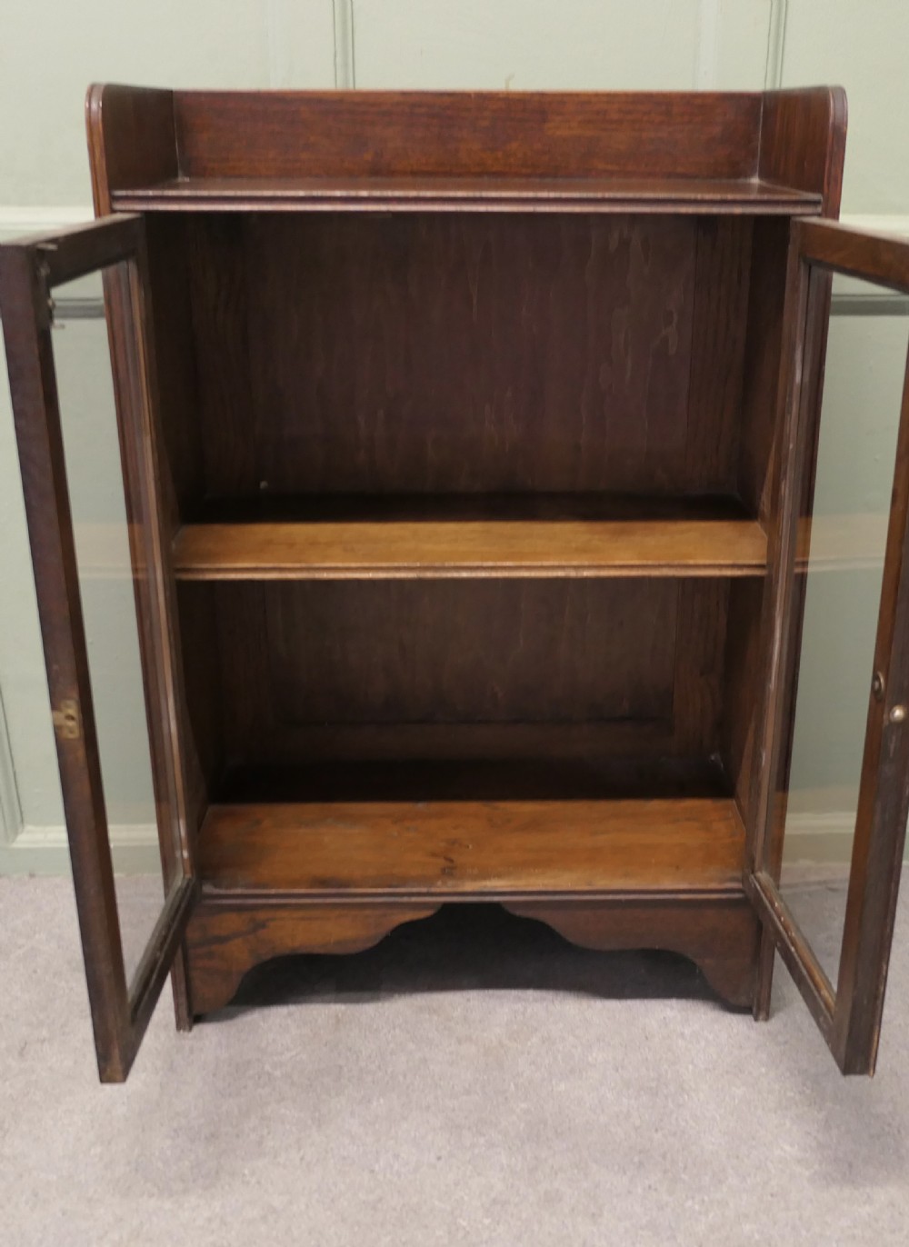 A small dark oak glazed bookcase featuring twin glass panel doors, a raised top gallery, and a shaped lower apron. View IV