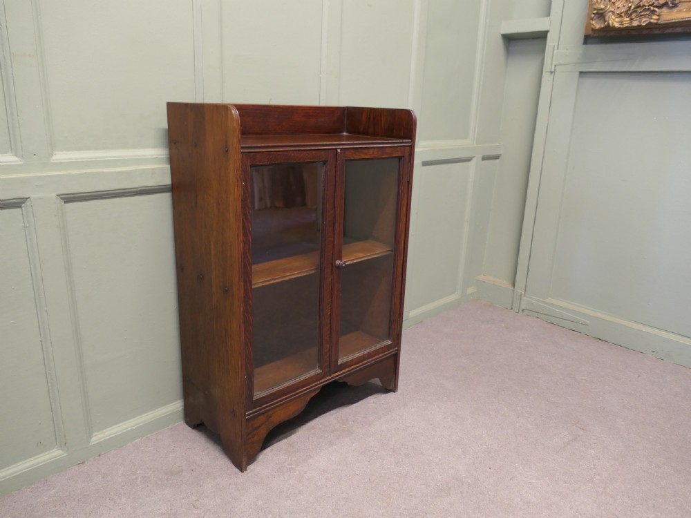 A small dark oak glazed bookcase featuring twin glass panel doors, a raised top gallery, and a shaped lower apron. View V