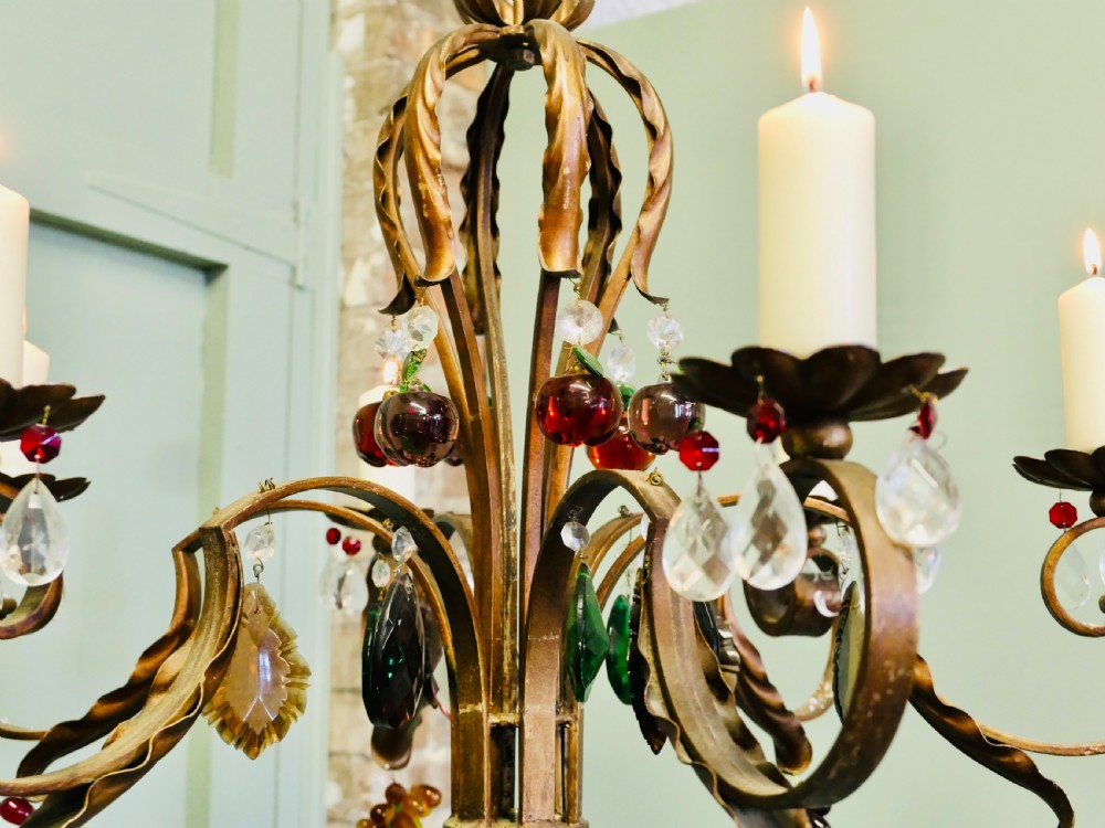 A six-branch wrought iron chandelier decorated with leaf motifs and suspended coloured glass fruit drops, including grapes, apples, and pears. View VI