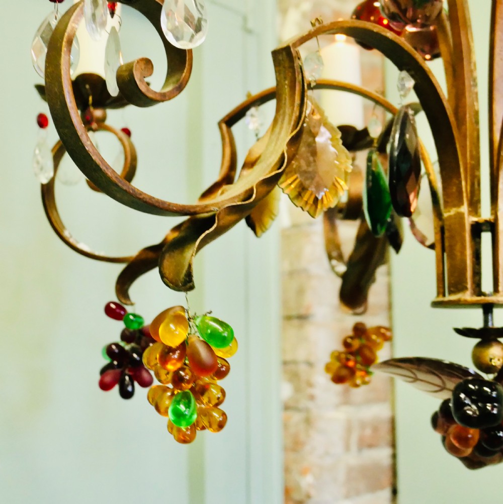 A six-branch wrought iron chandelier decorated with leaf motifs and suspended coloured glass fruit drops, including grapes, apples, and pears. View IV