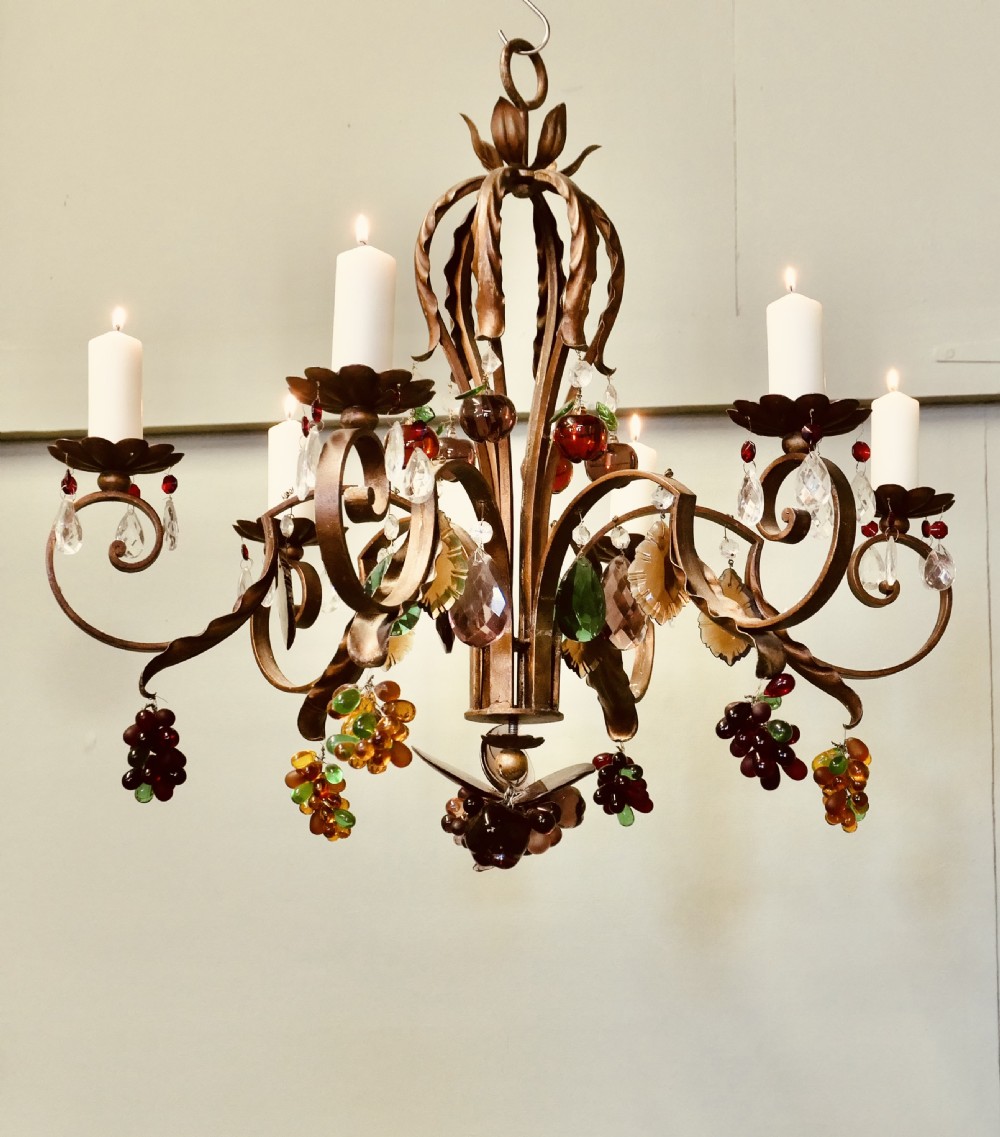 A six-branch wrought iron chandelier decorated with leaf motifs and suspended coloured glass fruit drops, including grapes, apples, and pears.